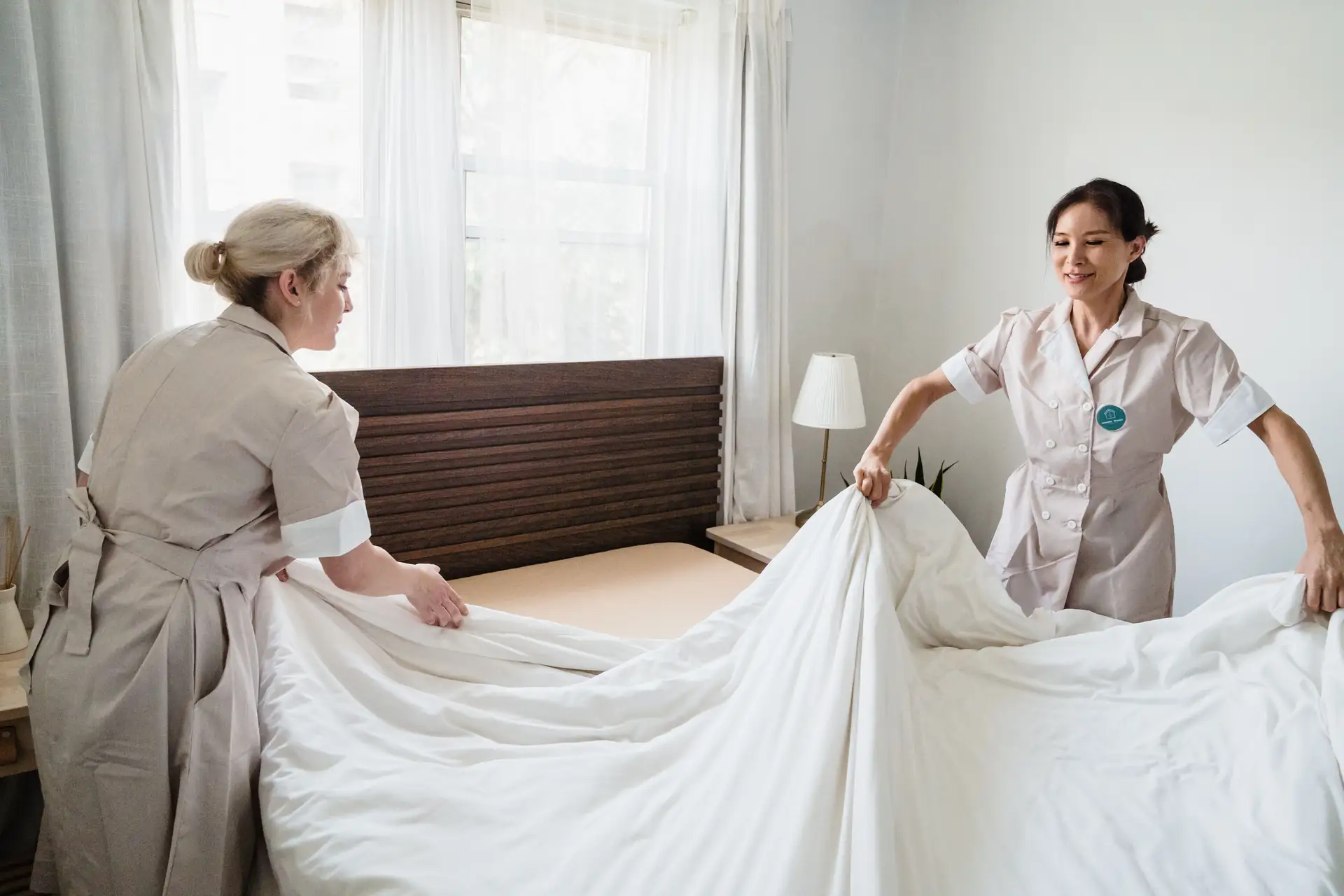 Duas camareiras estendendo um lençol branco sobre a cama de um quarto claro